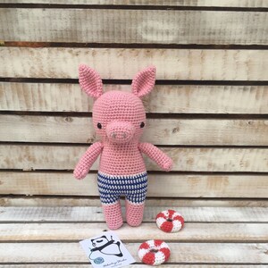 May include: Pink crocheted pig wearing blue and white striped shorts. The pig is standing on a wooden surface with two red and white crocheted life preservers.