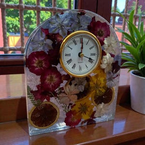 May include: A decorative quartz clock with a gold-tone bezel and Roman numerals. The clock face is white. The clear resin base is embedded with dried flowers and orange slices, creating a unique, floral design.