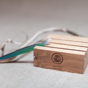 May include: A wooden stand with four slots for holding items. The stand is made of light brown wood and has the words "NATIVE CIRCLES" engraved on the bottom. A blue and green feather is resting on top of the stand.