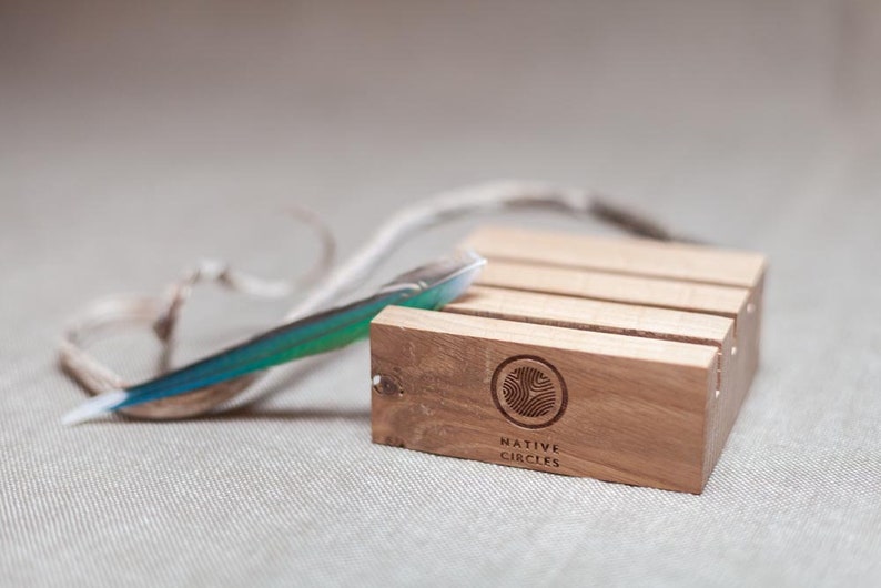 May include: A wooden stand with four slots for holding items. The stand is made of light brown wood and has the words "NATIVE CIRCLES" engraved on the bottom. A blue and green feather is resting on top of the stand.