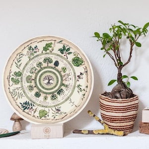 May include: A round, cream-colored plate with a circular design of green trees and leaves. The plate is on a wooden stand. There is a small potted plant with green leaves and a brown woven basket in the background. The plate has the text "Nature's Cycles" printed on it.