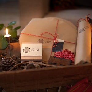 May include: A rustic wooden basket filled with brown paper wrapped gifts, a candle, and pine cones. The gifts are tied with red string and have the logo "NATIVE CIRCLES" printed on them.