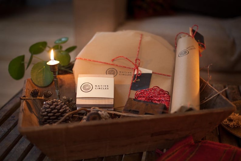 May include: A rustic wooden basket filled with brown paper wrapped gifts, a candle, and pine cones. The gifts are tied with red string and have the logo "NATIVE CIRCLES" printed on them.