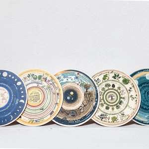 May include: Five round, colorful, illustrated paper plates with different designs. The plates feature a moon phase calendar, a tree of life, a seasonal calendar, a winter solstice calendar, and a garden calendar.
