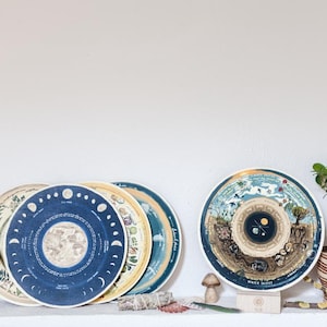 May include: A set of five round, decorative plates with blue and gold designs. The plates feature illustrations of the moon phases, the earth, and other celestial bodies. The plates are arranged in a stack on a white shelf with a small potted plant, a bundle of dried herbs, and a mushroom figurine.