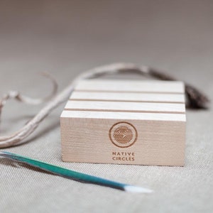 May include: A wooden display stand with four slots for rings or other small items. The stand is made of light wood and has a natural finish. The stand has the words "NATIVE CIRCLES" engraved on the front.