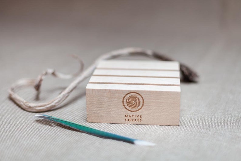 May include: A wooden display stand with four slots for rings or other small items. The stand is made of light wood and has a natural finish. The stand has the words "NATIVE CIRCLES" engraved on the front.