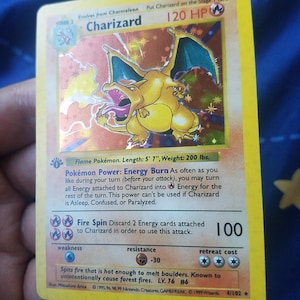May include: A Charizard Pokémon trading card. The card features a gold and orange Charizard illustration with blue wings. The card has text including "120 HP", "Fire Spin", and "LV. 76 #6". The card is 5' 7" long and weighs 200 lbs.