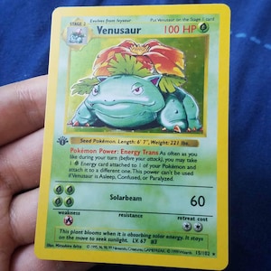Peut inclure: Une carte à collectionner rectangulaire représentant le personnage vert Florizarre avec une grande fleur rouge sur le dos. La carte affiche 100 PV, le texte Pokémon Power: Energy Trans, Lance-Soleil 60 et un tampon 1st Edition sur bordure jaune.