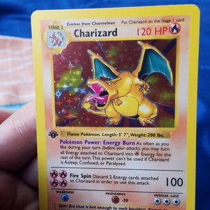 May include: A holographic Charizard trading card featuring a yellow border, 120 HP, and a Stage 2 evolution icon. The card displays the Flame Pokémon with a fire-breathing illustration, Energy Burn power text, and a 1st Edition stamp on the left side.