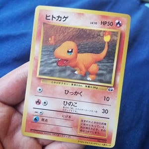 May include: A vintage Pokémon trading card featuring Charmander, an orange lizard-like creature with a flame on its tail. The card has Japanese text and a HP50 rating. The card is held in a hand.