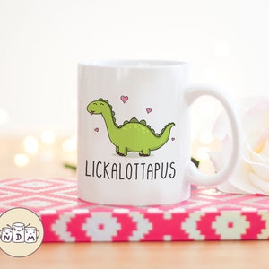 以下が含まれることがあります： 白いセラミックマグカップ。緑色の漫画の恐竜と「LICKALOTTAPUS」の文字が描かれています。 恐竜は舌を出しており、ピンクのハートに囲まれています。 マグカップはピンクと白の模様の表面に置かれています。