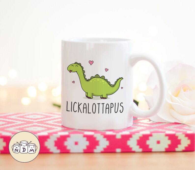以下が含まれることがあります： 白いセラミックマグカップ。緑色の漫画の恐竜と「LICKALOTTAPUS」の文字が描かれています。 恐竜は舌を出しており、ピンクのハートに囲まれています。 マグカップはピンクと白の模様の表面に置かれています。