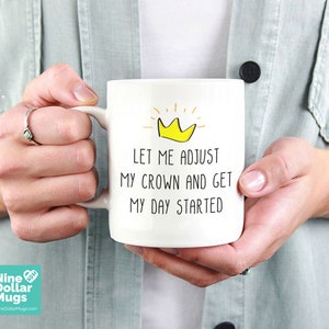 May include: White ceramic mug with the text "LET ME ADJUST MY CROWN AND GET MY DAY STARTED" and a yellow crown graphic. The mug is held by a person. The mug has a large handle.