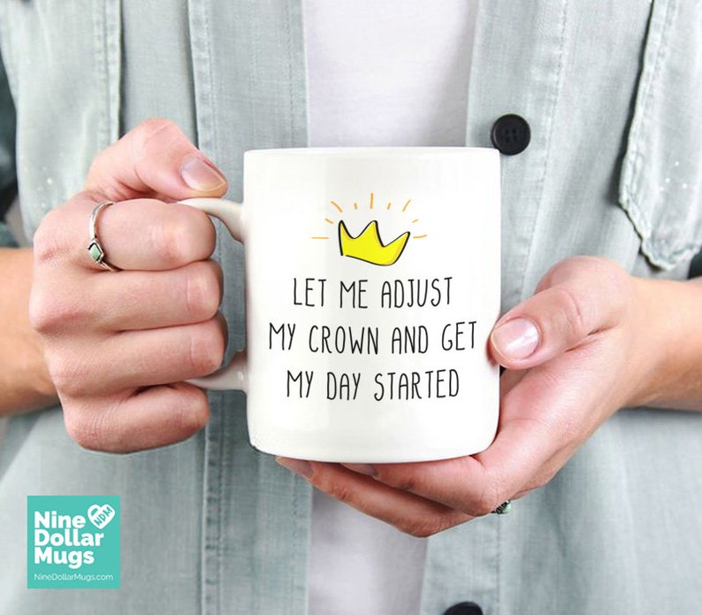May include: White ceramic mug with the text "LET ME ADJUST MY CROWN AND GET MY DAY STARTED" and a yellow crown graphic. The mug is held by a person. The mug has a large handle.