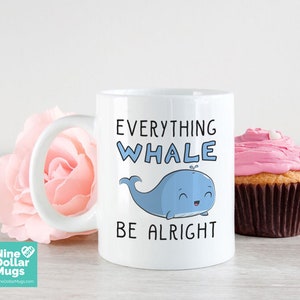 K&ouml;nnte beinhalten: Wei&szlig;e Keramik-Tasse mit dem Text "Everything Whale Be Alright" und einer blauen Cartoon-Wal-Illustration. Eine rosa Blume und ein Cupcake mit rosa Zuckerguss sind im Hintergrund. Die Tasse ist von Nine Dollar Mugs.