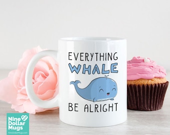 May include: White ceramic mug with the text "Everything Whale Be Alright" and a cartoon whale illustration in blue. A pink flower and a cupcake with pink frosting are in the background. The mug is from Nine Dollar Mugs.