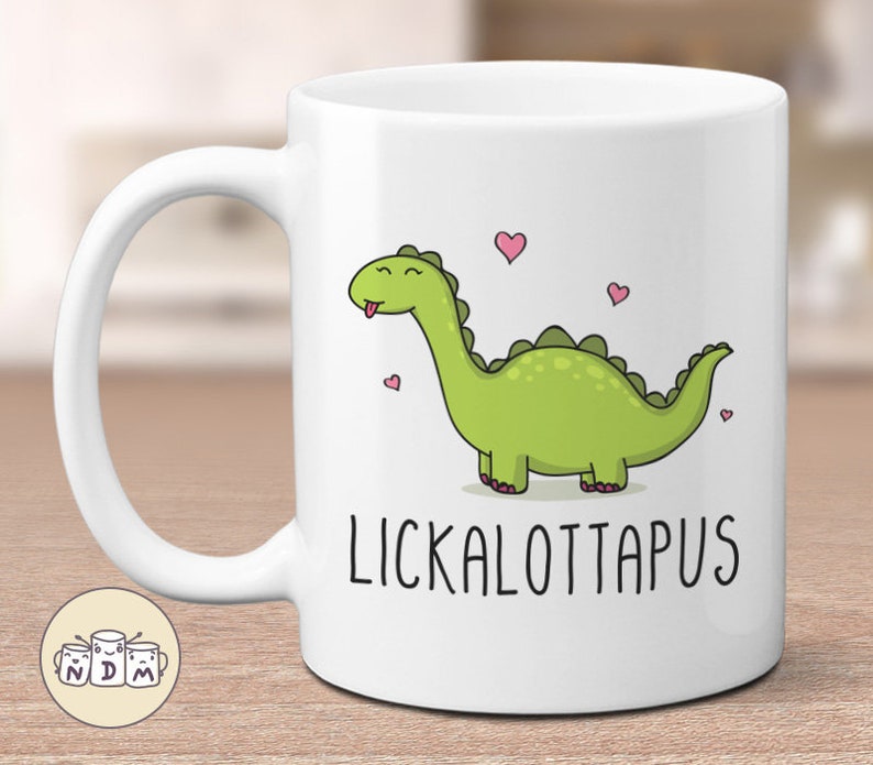 以下が含まれることがあります： 白いセラミックマグカップ。緑色の漫画の恐竜と「LICKALOTTAPUS」の文字が描かれています。恐竜は長い首を持ち、ピンクのハートに囲まれています。楽しくて気まぐれなデザイン。コーヒーや紅茶に最適です。