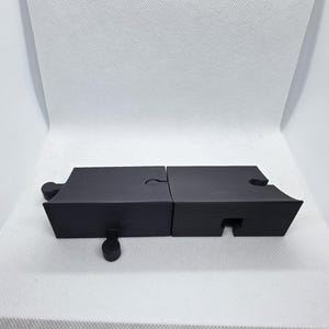 Pode incluir: Duas pe&ccedil;as de quebra-cabe&ccedil;a pretas interligadas, formando uma forma retangular. As pe&ccedil;as t&ecirc;m um acabamento fosco e est&atilde;o sobre um fundo branco. Uma pe&ccedil;a tem um pequeno pino cil&iacute;ndrico.