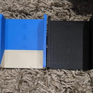 Pode incluir: Dois recipientes retangulares abertos. Um &eacute; azul com base bege, o outro &eacute; preto. Ambos t&ecirc;m tr&ecirc;s recortes retangulares na frente. Os recipientes est&atilde;o sobre uma superf&iacute;cie texturizada marrom claro.