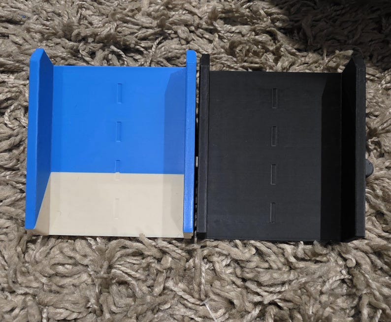 Pode incluir: Dois recipientes retangulares abertos. Um &eacute; azul com base bege, o outro &eacute; preto. Ambos t&ecirc;m tr&ecirc;s recortes retangulares na frente. Os recipientes est&atilde;o sobre uma superf&iacute;cie texturizada marrom claro.