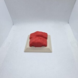 Pode incluir: Dois modelos de carros vermelhos s&atilde;o exibidos em uma base retangular branca. Os carros s&atilde;o de cor vermelha s&oacute;lida e parecem r&eacute;plicas em miniatura. A base &eacute; de cor bege claro.