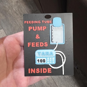 Op de afbeelding: Een zwart rechthoekig bord met de woorden "FEEDING TUBE PUMP & FEEDS INSIDE" in rode en witte tekst. Het bord toont een blauw-witte illustratie van een voedingspomp met het woord "TARA" en het nummer "100".