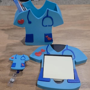 May include: A collection of medical-themed desk accessories. Includes a light blue pen holder, a notepad holder, and a badge reel, all shaped like medical scrubs. Each item features a stethoscope design and red heart accents.