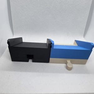 Pode incluir: Um brinquedo de madeira geom&eacute;trico multicolorido. O brinquedo &eacute; principalmente preto e azul, com uma base bege. Tem uma forma retangular com um pequeno pino bege saliente. O brinquedo &eacute; projetado com uma ranhura e uma se&ccedil;&atilde;o elevada.