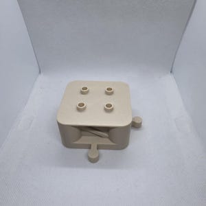 Pode incluir: Objeto bege impresso em 3D em forma de cubo, com quatro orif&iacute;cios cil&iacute;ndricos na parte superior e uma protuber&acirc;ncia em forma de al&ccedil;a na lateral. O objeto tem um design minimalista e parece ser um item funcional.