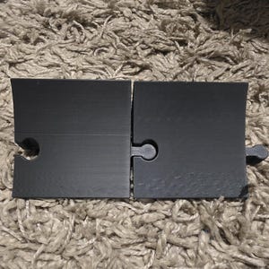Pode incluir: Duas pe&ccedil;as de quebra-cabe&ccedil;a pretas foscas, interligadas, com um conector em forma de buraco de fechadura. As pe&ccedil;as retangulares t&ecirc;m uma borda superior ligeiramente angulada. O fundo &eacute; um tapete texturizado castanho claro.