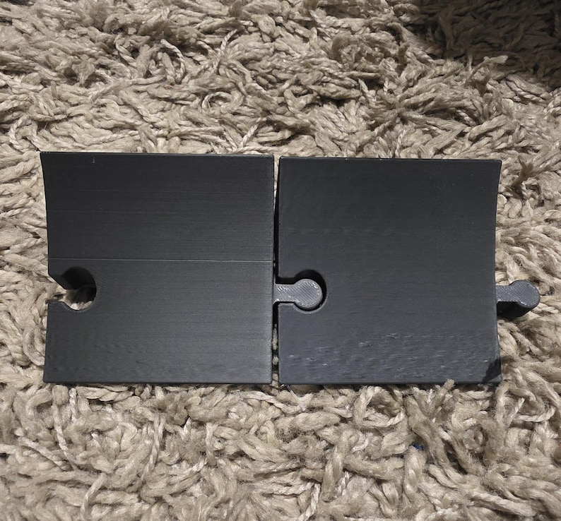 Pode incluir: Duas pe&ccedil;as de quebra-cabe&ccedil;a pretas foscas, interligadas, com um conector em forma de buraco de fechadura. As pe&ccedil;as retangulares t&ecirc;m uma borda superior ligeiramente angulada. O fundo &eacute; um tapete texturizado castanho claro.