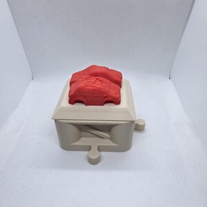 Pode incluir: Uma caixa bege com um objeto vermelho em forma de carro em cima. A caixa tem uma forma retangular com um topo plano e um design decorativo na frente. O objeto em forma de carro &eacute; de cor vermelha brilhante e parece ser um brinquedo ou modelo.