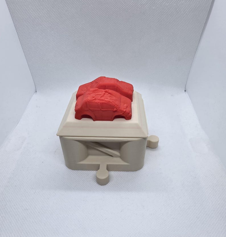Pode incluir: Uma caixa bege com um objeto vermelho em forma de carro em cima. A caixa tem uma forma retangular com um topo plano e um design decorativo na frente. O objeto em forma de carro &eacute; de cor vermelha brilhante e parece ser um brinquedo ou modelo.