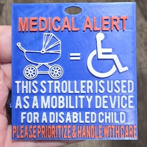 May include: A square blue sign with orange and white raised text and graphics. It features a white stroller icon, an equals sign, and a white wheelchair icon. The text reads: MEDICAL ALERT, THIS STROLLER IS USED AS A MOBILITY DEVICE FOR A DISABLED CHILD, PLEASE PRIORITIZE & HANDLE WITH CARE.