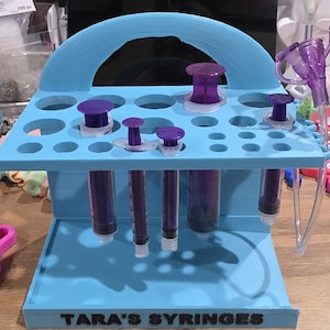 以下が含まれることがあります： ベースに「TARA'S SYRINGES」の文字が入った水色の注射器ホルダー。ホルダーには複数の穴があり、紫色の注射器と紫色の注射器キャップがいくつか入っています。紫色の医療機器が側面に付いています。