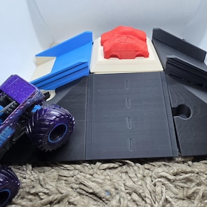 Pode incluir: Um brinquedo de caminh&atilde;o monstro roxo e azul com pneus grandes est&aacute; posicionado ao lado de um conjunto de pistas impresso em 3D. O conjunto de pistas inclui uma rampa azul, um carro vermelho e pe&ccedil;as de pista pretas. O conjunto &eacute; projetado para brincar com carrinhos.