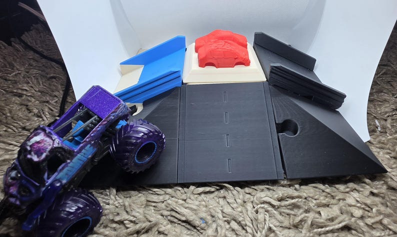 Pode incluir: Um brinquedo de caminh&atilde;o monstro roxo e azul com pneus grandes est&aacute; posicionado ao lado de um conjunto de pistas impresso em 3D. O conjunto de pistas inclui uma rampa azul, um carro vermelho e pe&ccedil;as de pista pretas. O conjunto &eacute; projetado para brincar com carrinhos.