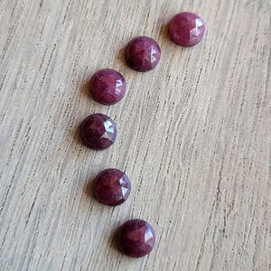 May include: Six small, round, faceted gemstones in a deep magenta-red color. Each stone features a geometric, multi-faceted surface that reflects light, arranged in a gentle, slightly curved vertical line formation on a light wood grain surface.