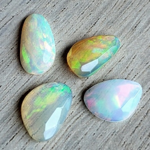 May include: Four faceted opal gemstones with iridescent color play. The opals display a range of colors, including white, green, yellow, and pink. The gemstones are set against a gray textured background.