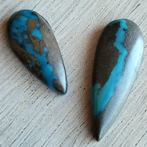 May include: Two teardrop-shaped gemstones with a mix of blue, brown, and gray hues. The stones have a polished surface and a natural, organic appearance. The gemstones are set against a light-colored, textured background.