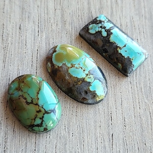 May include: Three polished turquoise cabochons featuring natural matrix patterns. One stone is oval-shaped with dark brown veining, another is a larger oval with green and blue hues, and the third is a rectangular piece with dark matrix.