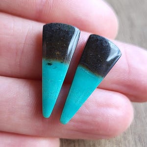 May include: Two turquoise and black triangular earrings. The top portion of each earring is black, transitioning to a brown band, and then a vibrant turquoise color at the bottom. The earrings are displayed on a wooden surface.