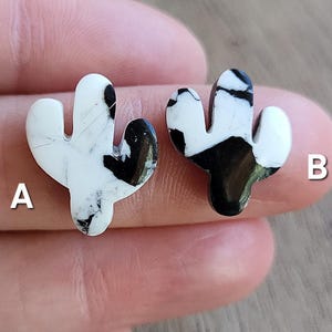 May include: Two cactus-shaped stud earrings. Each earring is white with black marbling. The earrings are small and have a three-dimensional design. The earrings are shown on a person's finger.