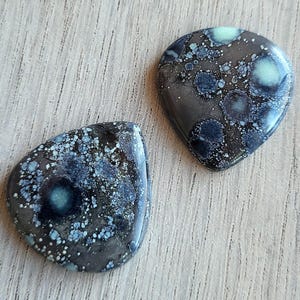 May include: Two polished, teardrop-shaped stones with a speckled pattern. The stones feature a dark gray base color with blue, teal, and white speckles and circular patterns. The stones are displayed on a light wood surface.