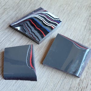 May include: Three decorative coasters with a layered design. One coaster is rectangular with a pattern of red, blue, white, and black stripes. The other two coasters are gray with a red and white stripe design on the edge.