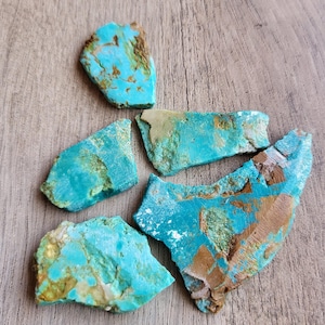 May include: A collection of raw turquoise stones with a vibrant blue-green hue and brown matrix. The stones vary in size and shape, displaying natural textures and patterns. They are arranged on a light brown wooden surface.
