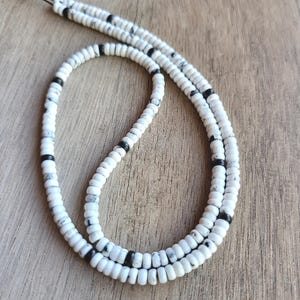 May include: A long necklace composed of small, round, white beads with black marble-like veining, interspersed with solid black beads at regular intervals. The beads are strung on a thin black cord that is visible at the top of the necklace.