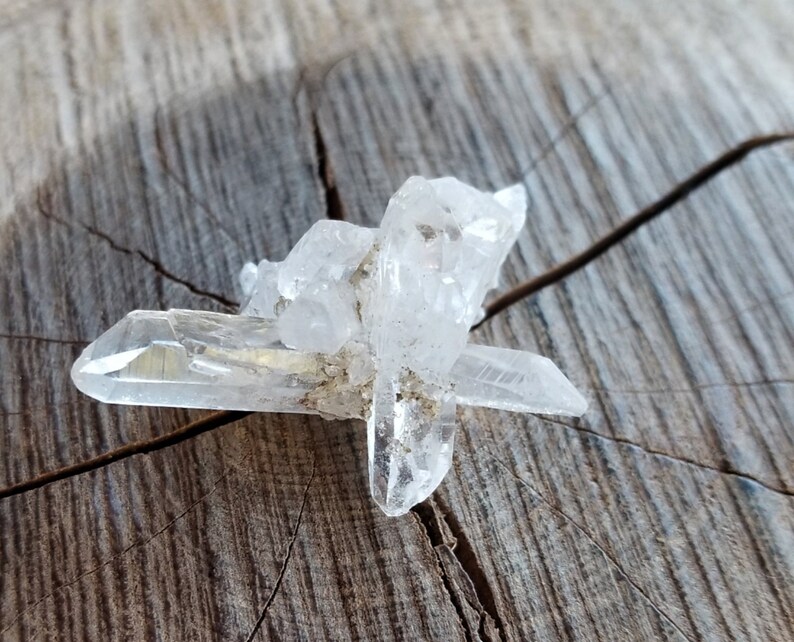 May include: A cluster of clear quartz crystals with a pointed shape, resting on a piece of weathered wood.