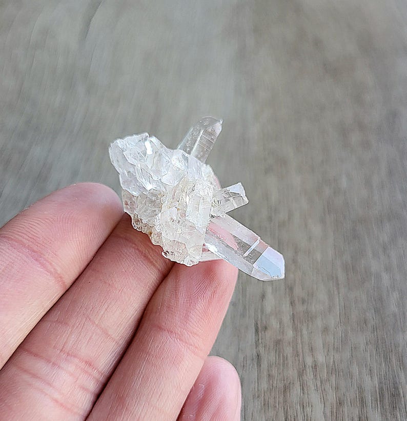 May include: A small cluster of clear quartz crystals with a rainbow iridescent effect. The crystals are clustered together in a natural formation.